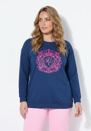 Vrouw met golvend blond haar die een marineblauwe trui draagt met een roze sierlijk wapenschild en lichtroze broek tegen een witte achtergrond.