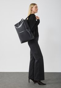 Mochila negra brillante con forma rectangular, que cuenta con dos correas ajustables y un asa en la parte superior, exhibida sobre un fondo neutro.