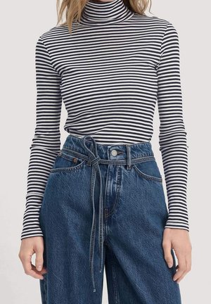Femme portant un col roulé à manches longues rayé noir et blanc, rentré dans un jean bleu taille haute avec une ceinture nouée.