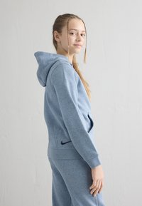 Adolescentă cu șuvițe împletite, purtând un hanorac albastru și pantaloni de trening asortați, stând în profil în fața unui perete alb simplu.