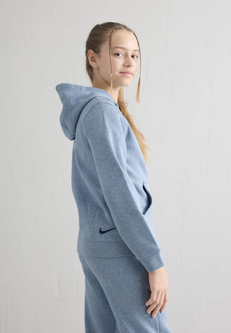 Adolescentă cu șuvițe împletite, purtând un hanorac albastru și pantaloni de trening asortați, stând în profil în fața unui perete alb simplu.