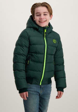 Boy smiles while wearing a green puffer jacket with a bright yellow zipper and blue jeans, standing against a plain light background.
