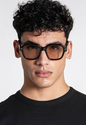 Ung mand med krøllet mørkt hår, iført store sorte briller med ravfarvede glas og en lille guldring i øret, klædt i en sort ribstrikket skjorte.