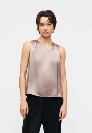 Jonge vrouw met kort haar, gekleed in een mouwloos satijnen taupekleurig topje en zwarte geplooide broek, staand tegen een effen witte achtergrond.