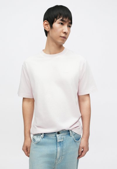 Ροζ βαμβακερό t-shirt με στρογγυλή λαιμόκοψη και κοντά μανίκια, με διακριτικές λεπτομέρειες λογότυπου. Συνδυάζεται με ανοιχτό μπλε τζιν παντελόνι.