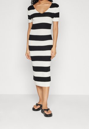 Femme portant une robe tricotée à rayures horizontales noires et blanches à manches courtes et des sandales plateformes noires, debout contre un fond uni.