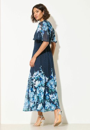 Frau trägt ein marineblaues Maxikleid mit großem hellblauem Blumenmuster und flatternden Ärmeln, kombiniert mit silbernen Sandalen mit hohen Absätzen, seitlich stehend.