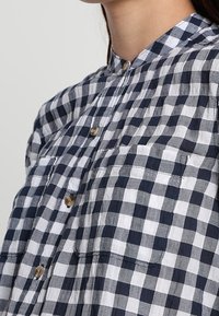 Camisa de botones en patrón de cuadros gales en navy y blanco, con un collar clásico, dos bolsillos en el pecho y botones marrones.