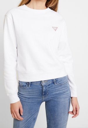 Woman wearing a white Guess sweatshirt with a small red logo on the chest and blue jeans, standing with her hands relaxed by her sides.