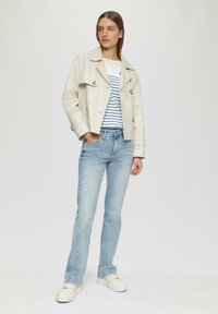 Veste courte beige clair avec un boutonnage croisé sur une chemise rayée blanche et bleue, assortie à un jean délavé bleu et des baskets blanches.