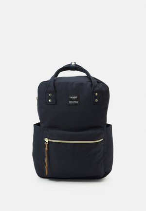 Mochila de lona negra con forma rectangular, dos asas superiores, bolsillo frontal con cremallera, acentos de hardware dorado y una pequeña etiqueta de logo.