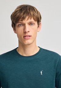 Maglietta a maniche lunghe di un verde petrolio scuro con scollo tondeggiante, che presenta un piccolo logo ricamato bianco di un golfista sul lato sinistro del petto. Tessuto morbido.