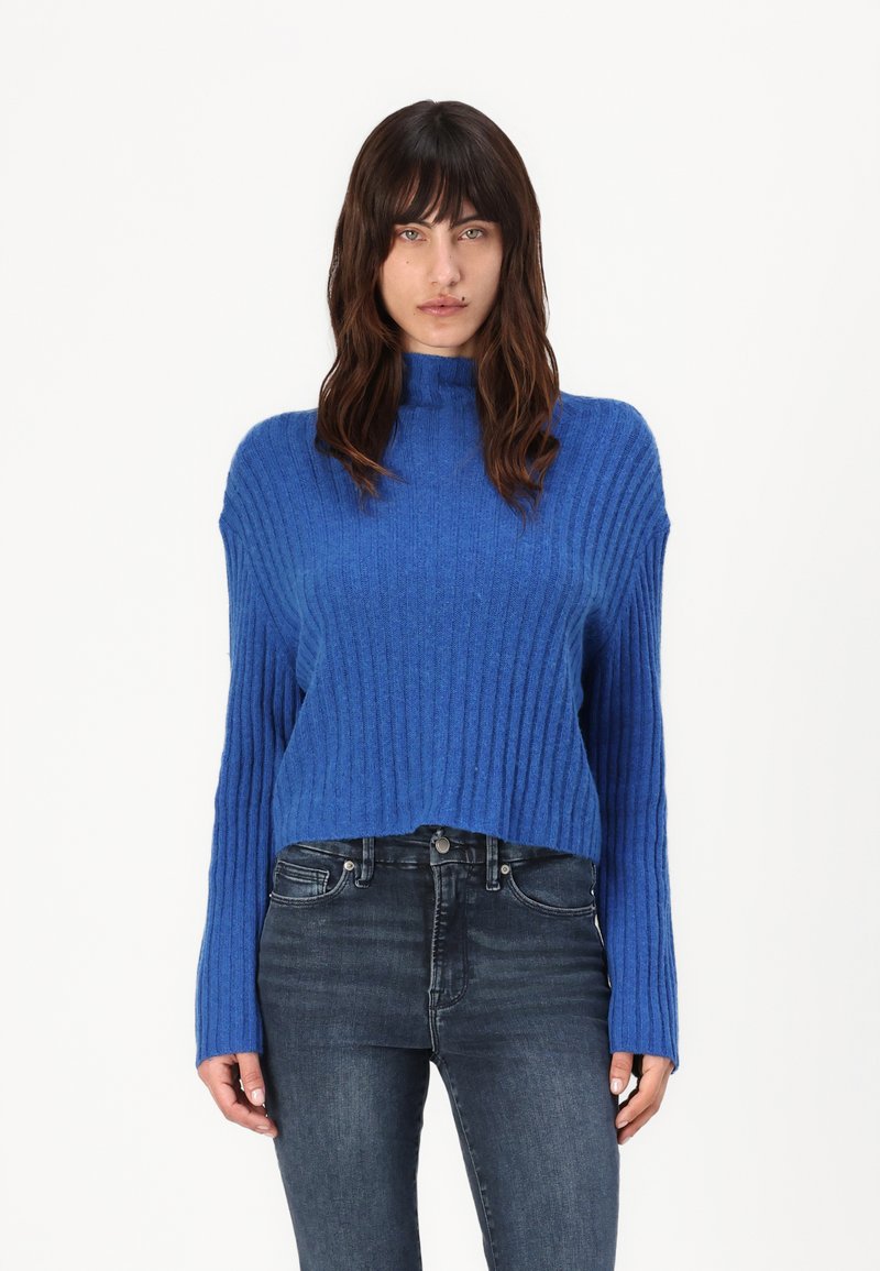 Strikket blå genser med høy hals og ribbestrikket tekstur, med lange ermer og kort lengde, kombinert med mørke jeans.
