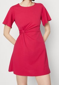Dopasowana sukienka z krótkim rękawem w jaskrawym różu. Posiada detal w formie węzła w talii oraz rozkloszowany dół. Wykonana z gładkiego, elastycznego materiału.