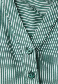 Grønt stof med lodrette striber og en blød tekstur, der har to teal knapper og en V-hals design. Bemærkelsesværdige syninger kan ses.