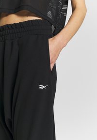 Schwarze Jogginghose mit elastischem Bund, seitlichen Taschen und einem weißen Logo am unteren Bein. Das Material wirkt weich und strukturiert.
