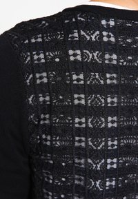 Svart stickad topp med ett mönstrat spetsrygg, dekorerad med horisontella ränder och blommotiv. Den strukturerade tyget kontrasterar med den släta stickningen.