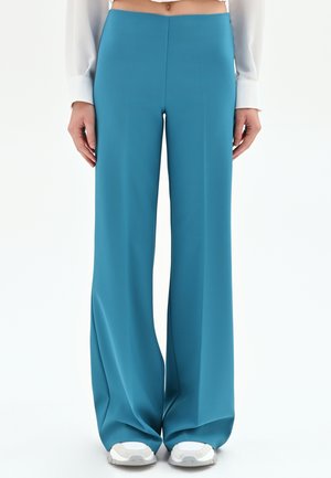 Femme portant un pantalon large taille haute bleu sarcelle, associé à des baskets blanches et un haut blanc à manches longues, debout devant un fond blanc.