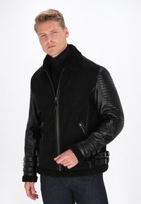 Veste aviateur noire avec col en peau de mouton, manches en cuir, poches zippées, texture côtelée sur les manches et accents à boucle à la taille.