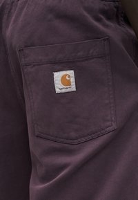 Κοντινό πλάνο μωβ τσέπης υφάσματος με ραμμένη στο κέντρο της τσέπης ένα μπαλώμα με το λογότυπο Carhartt.