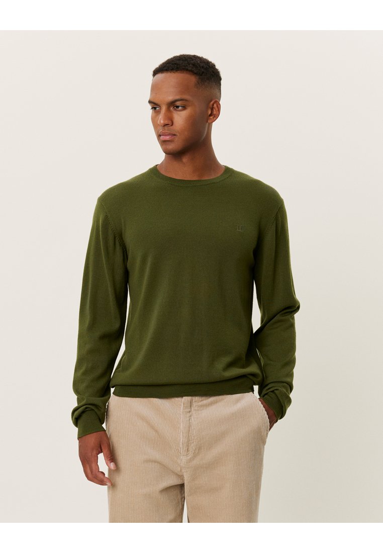 Pulover verde închis dintr-un material tricotat, cu mâneci lungi, guler rotund și un detaliu subtil al logo-ului pe piept. Purtați cu pantaloni crem deschis.