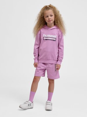 Ein junges Mädchen mit lockigem Haar steht, trägt einen pinken Kapuzenpullover und passende Shorts, pinke Socken und weiße Sneakers vor einem einfarbigen Hintergrund.