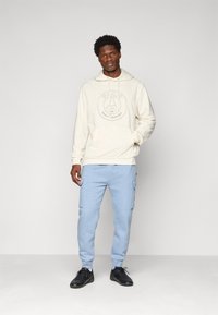Young man standing wearing beige Paris Saint-Germain hoodie, light blue cargo sweatpants, and black sneakers against plain gray background.