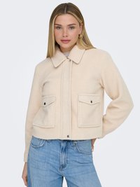 Ung kvinde med glat blondt hår iført en cremefarvet uldjakke med klaplommer og lyseblå jeans mod en ensfarvet baggrund.