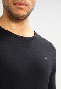 Pull en tricot noir avec un design en V, arborant un petit logo coloré sur la poitrine et une texture côtelée au niveau de l'encolure et des poignets.