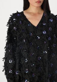 Blusa preta com estampa floral, apresentando um tecido texturizado, decote em V e numerosas aplicações de flores em paetês multicoloridos por toda a peça.