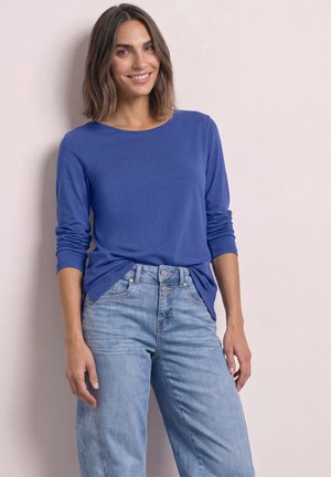 Smilende kvinde med skulderlangt brunt hår iført en langærmet blå skjorte og lyseblå højtaljede jeans mod en ensfarvet baggrund.