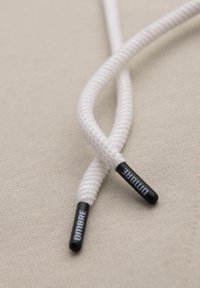 Lacci bianchi intrecciati con aglette nere che presentano il testo "DIMBRE" e "DUNBARE." Lo sfondo è una superficie tessile beige chiaro.