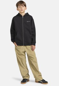 Sudadera con cremallera negra con logotipo, bolsillos frontales, combinada con pantalones beige holgados y zapatillas negras con suelas marrones. Textura de tela suave.