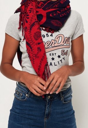 Personne portant un t-shirt graphique gris clair, un jean bleu et une écharpe rouge et marine à motifs enroulée autour du cou, les mains ajustant les vêtements.