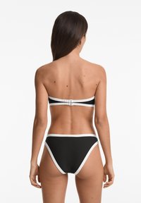 Črni bikini komplet, ki vključuje bandeau zgornji del z belimi poudarki in zaponko, v kombinaciji z visokim pasom. Gladka tekstura.