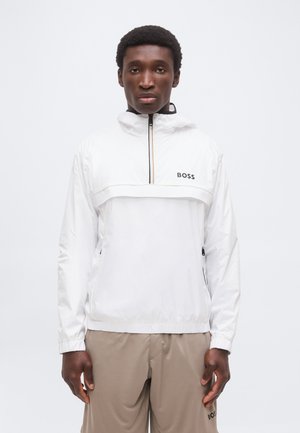 Άνδρας που φοράει λευκό μπουφάν BOSS με φερμουάρ και κουκούλα και μπεζ παντελόνια, στέκεται μπροστά σε ένα απλό λευκό φόντο.