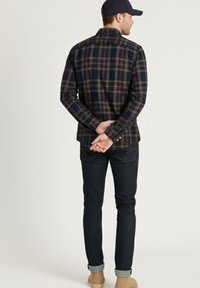 Camisa de franela a cuadros en tonos oscuros, de manga larga, con cuello abotonado, combinada con jeans oscuros y botas marrones, vista desde atrás.