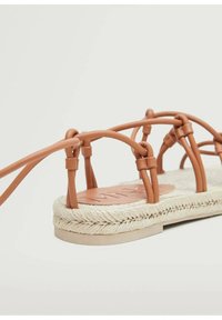 Sandale marron à brides avec semelle en jute tressée et semelle plate en caoutchouc beige sur fond blanc.