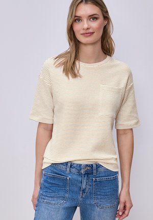 Kvinde iført en beige kortærmet t-shirt med striber og brystlomme samt højtsiddende blå cowboybukser mod en ensfarvet baggrund.