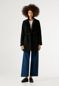 Melnās dubultā aizdare mētelis ar plašām atlokām, kopā ar ziliem plašām kājām džinsiem un melnām kurpēm, ar tekstūras rakstiem.