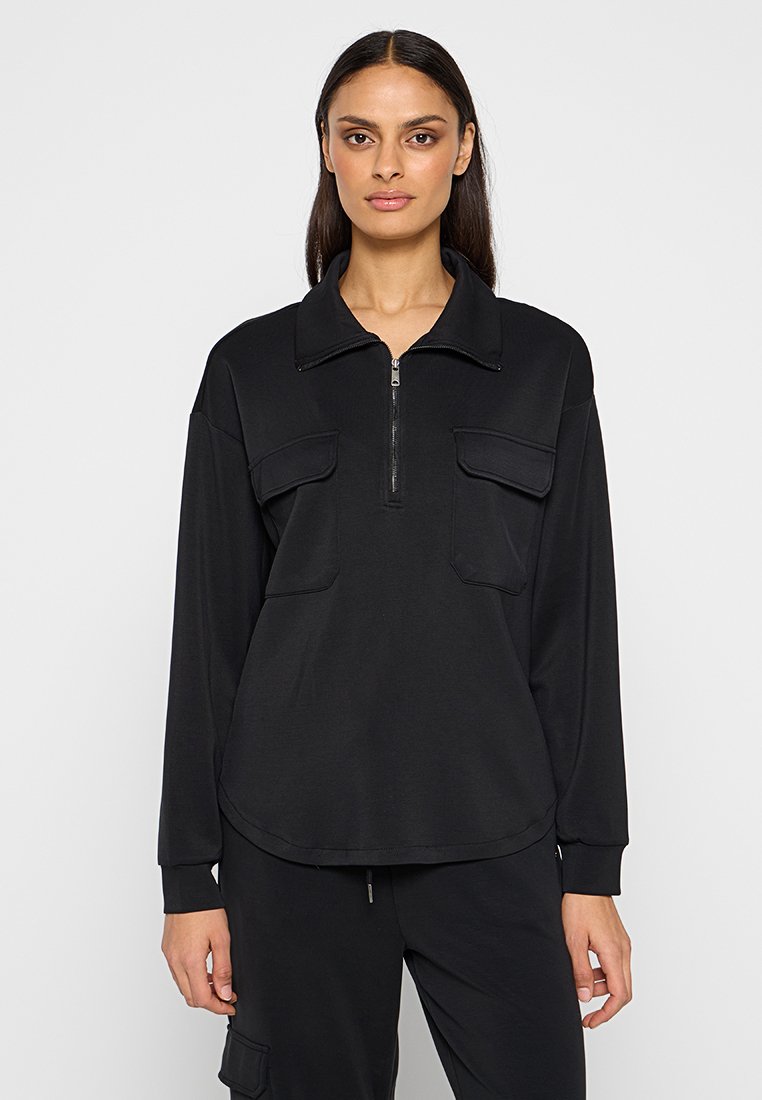 Schwarzer Pullover mit langen Ärmeln, der einen Reißverschlusskragen, zwei frontale Taschen und einen abgerundeten Saum aufweist, gefertigt aus einem weichen, glatten Stoff.