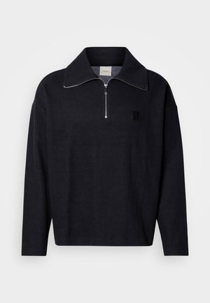 Pulover bleumarin închis cu guler înalt, fermoar frontal pe jumătate și un patch pătrat cu logo. Confecționat dintr-un material moale, texturat, cu mâneci lungi.