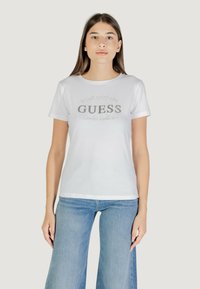 T-shirt in cotone bianco a maniche corte, con logo argentato "GUESS" e stampa "Established 1981". Tessuto liscio, vestibilità standard.