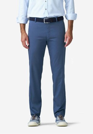 Uomo con camicia azzurra chiusa da bottoni, cintura blu scuro, pantaloni slim fit blu e sneakers casual bianche e blu, in piedi su sfondo bianco.