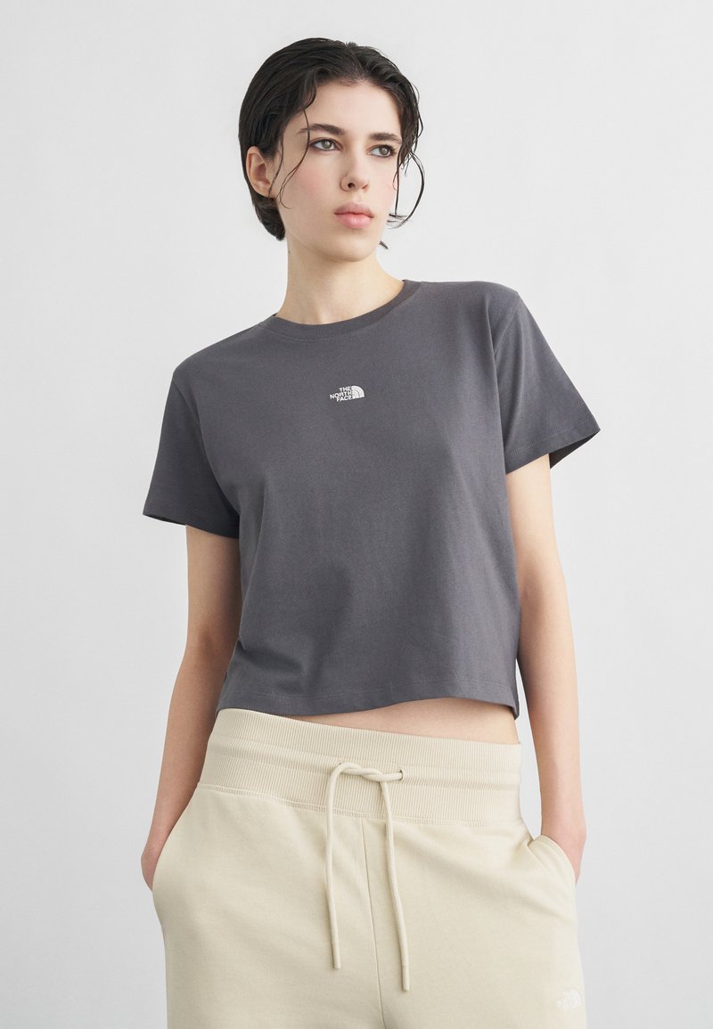 Νεαρό άτομο με κοντά σκούρα μαλλιά, φορώντας γκρι κοντομάνικο t-shirt North Face και μπεζ φόρμα με τα χέρια στις τσέπες.