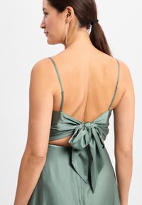 Grünes Satin-Kleid mit dünnen Trägern und einer großen Schleife auf der Rückseite. Der Stoff hat eine glatte Textur und zeigt einen Ausschnitt.