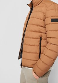 Giacca a vento marrone con sezioni imbottite orizzontali, colletto alto, zip nera e tasche laterali con eleganti dettagli neri.