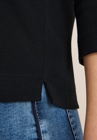 Musta puuvillapaita, jossa on pyöristetty helma ja sivuventtiilit. Kappaleen pinta on sileä, ja kauluksen sekä hihojen saumat ovat näkyvät.