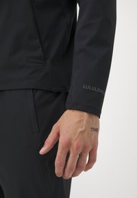 Schwarze wasserabweisende Jacke mit glatter Oberfläche, mit einem Lululemon-Logo auf dem �Ärmel. Hand mit sichtbar tätowiertem Handgelenk.