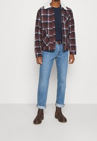 Chaqueta de franela a cuadros en burdeos, blanco y azul marino con cuello blanco, combinada con una camiseta azul marino de cuello redondo y vaqueros azul claro. Botas marrones.